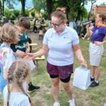 Na zdjęciu w parku kobieta w białej koszulce rozdaje dzieciom torby z prezentami. W tle ludzie siedzą na ławkach pod drzewami. Dzień jest pogodny z zielenią drzew i trawą.