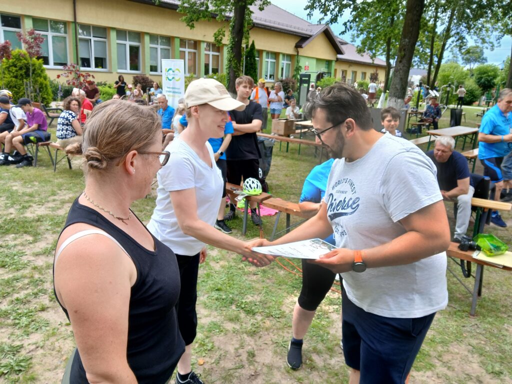 Na zdjęciu jest grupa ludzi na zewnątrz, jedna osoba wręcza certyfikat innej. Tło to budynek i drzewa, kolorystyka jest naturalna. Ludzie są ubrani w letnie ubrania.