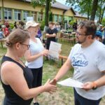 Na zdjęciu są dwie osoby podające sobie ręce na tle budynku. W tle są ludzie siedzący na ławkach. Zieleń dominuje w krajobrazie wokół.