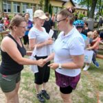 Grupa ludzi na zewnątrz, kobiety podają sobie ręce. Zielona trawa, stoliki piknikowe w tle. Relaksująca, przyjacielska atmosfera.