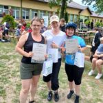 Trzy kobiety trzymają dyplomy na zielonej trawie. W tle budynek i ludzie na ławkach. Uśmiechają się, ubrani są sportowo.