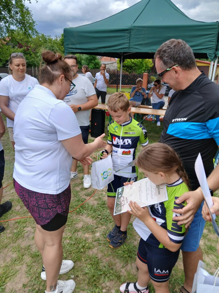Grupa osób w ogrodzie; dzieci w sportowych strojach z dyplomami, dorosły wręcza nagrody; zielony namiot w tle.