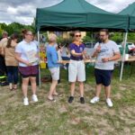 Na zdjęciu widać grupę osób pod zielonym namiotem na świeżym powietrzu. Jedna osoba trzyma mikrofon, inna trzyma dyplom. Tło to zielone krzewy i niebo z chmurami.