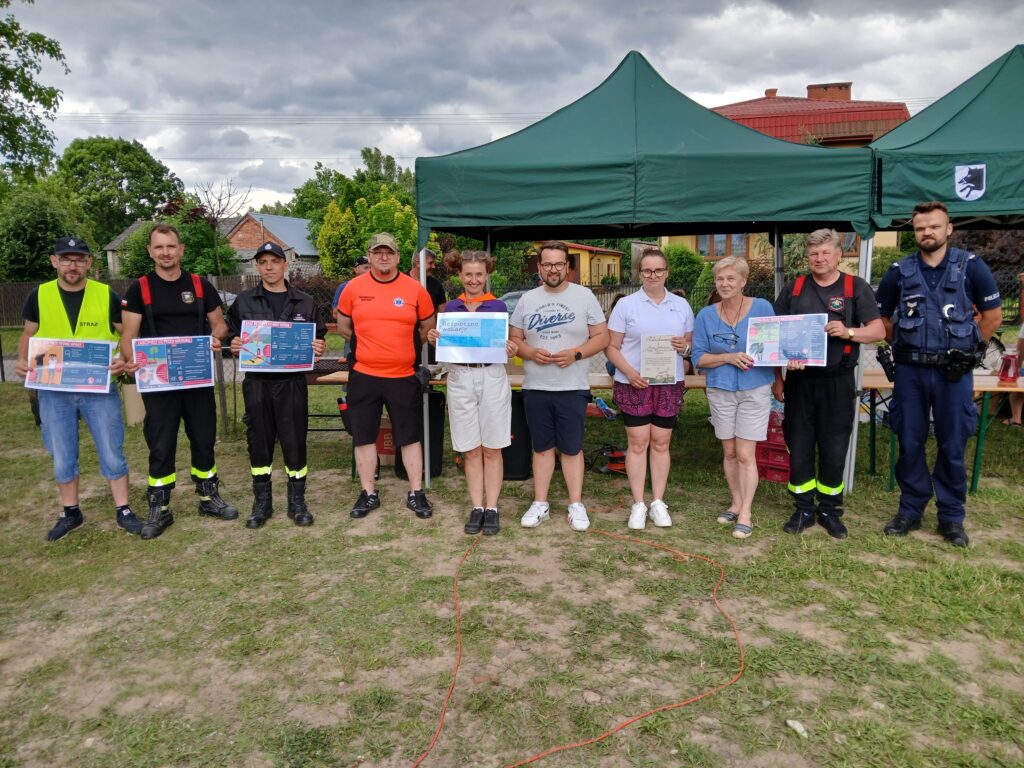 Grupa ludzi stoi na trawie przed zielonym namiotem. Każda osoba trzyma dyplom lub plakat. Wokół widoczne są drzewa i fragmenty budynków.