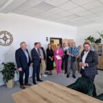 Grupa ludzi stoi w jasnym pomieszczeniu biurowym. Na ścianie zegar i rośliny w tle. Osoby są ubrane elegancko.