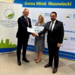 Na zdjęciu troje osób w formalnych strojach trzymających duży czek. Tło to baner Gminy Mińsk Mazowiecki w bieli i niebieskim. Po lewej widoczny jest roll-up z logo ekologicznej organizacji.