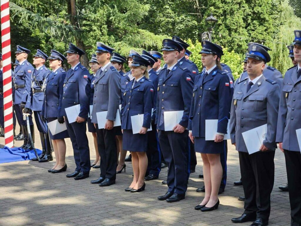 Grupa policjantów w granatowych i szarych mundurach stoi w szeregu. Każdy trzyma białą teczkę, tło to zielone drzewa. Pogodny dzień, cicha uroczystość na zewnątrz.