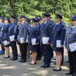 Grupa policjantów w granatowych i szarych mundurach stoi w szeregu. Każdy trzyma białą teczkę, tło to zielone drzewa. Pogodny dzień, cicha uroczystość na zewnątrz.