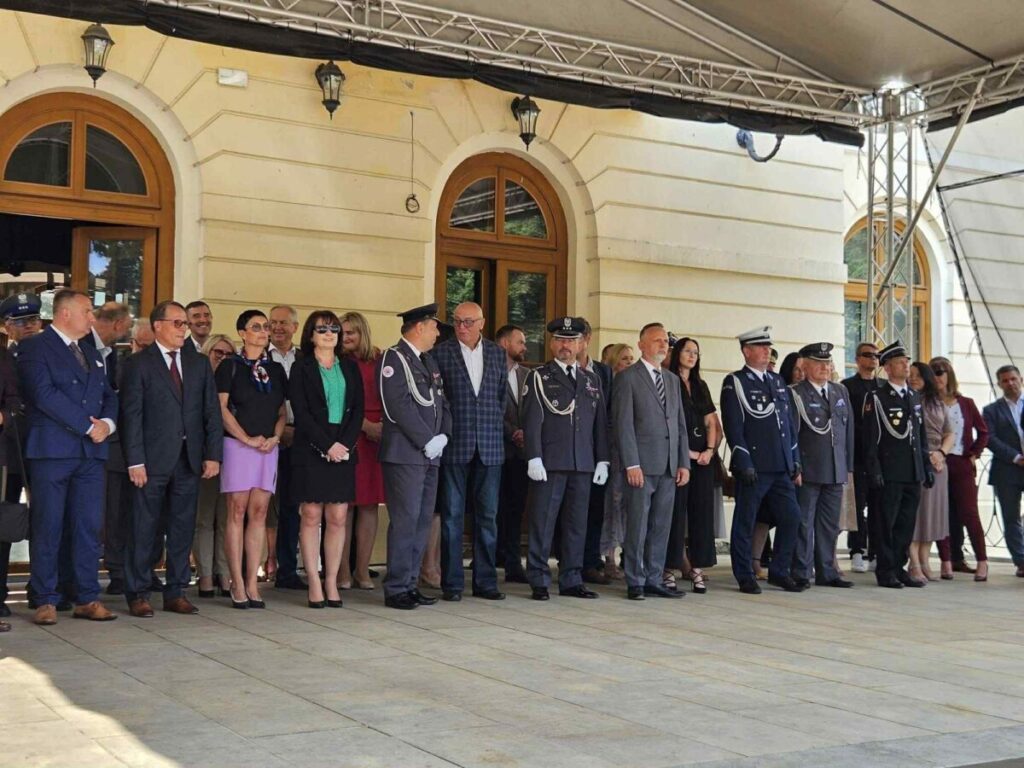 Grupa ludzi ubrana formalnie stoi przed jasnym budynkiem z łukowatymi oknami. W tle widać kremową ścianę i drewniane ramy okien. Niektórzy noszą mundury, inni garnitury i sukienki.