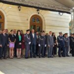 Grupa ludzi ubrana formalnie stoi przed jasnym budynkiem z łukowatymi oknami. W tle widać kremową ścianę i drewniane ramy okien. Niektórzy noszą mundury, inni garnitury i sukienki.