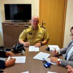 Trzech mężczyzn przy stole podpisuje dokumenty. Na ścianie telewizor, meble brązowe, ubrania w odcieniach czerni i beżu. Jasny, biurowy wystrój.