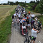 Rowerzyści na ścieżce wśród pól, niektórzy prowadzą rowery, tło zielonych drzew i niebo z chmurami.