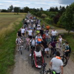 Grupa ludzi na rowerach jedzie po wiejskiej drodze. Jasne niebo z chmurami, zielone pola po bokach. Tłum na drodze odświętnie ubrany.