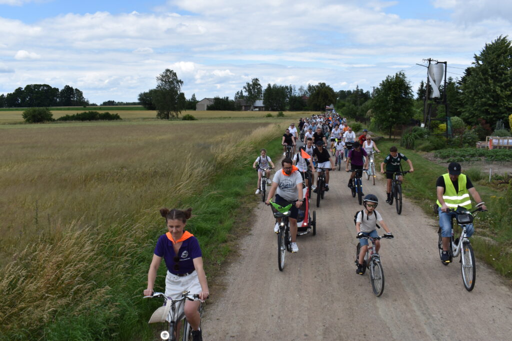 Grupa rowerzystów jedzie polną drogą wśród pól pod niebieskim niebem. W tle widoczne są drzewa i budynki. Uczestnicy mają kolorowe kamizelki i różnorodne stroje.