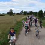 Grupa rowerzystów jedzie polną drogą wśród pól pod niebieskim niebem. W tle widoczne są drzewa i budynki. Uczestnicy mają kolorowe kamizelki i różnorodne stroje.