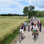 Duża grupa rowerzystów jedzie wzdłuż polnej drogi otoczonej zbożem. Krajobraz jest zielony, pod błękitnym niebem z chmurami. Tło stanowi las i pola.