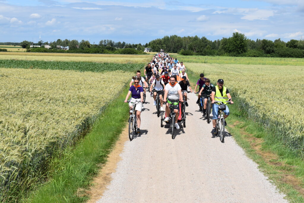 Grupa rowerzystów jedzie drogą polną przez zielone pola pod błękitnym niebem. Przyroda otacza trasę po obu stronach. Uczestnicy są różnorodnie ubrani.