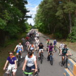 Grupa ludzi jedzie na rowerach wzdłuż asfaltowej drogi otoczonej zielenią drzew. Jest słoneczny dzień, w tle widać więcej uczestników. Po prawej stronie znajduje się chodnik i czerwona hydrantowa skrzynka.