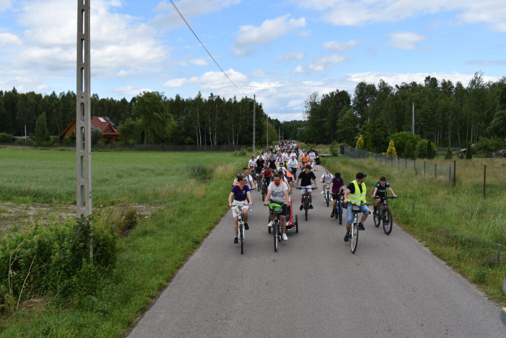 Grupa rowerzystów jedzie wzdłuż wiejskiej drogi. Otacza ich zielone pola i drzewa. Niebo jest częściowo pochmurne.
