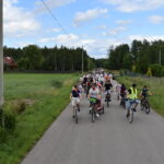 Grupa rowerzystów jedzie wzdłuż wiejskiej drogi. Otacza ich zielone pola i drzewa. Niebo jest częściowo pochmurne.