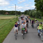 Grupa rowerzystów jedzie drogą polną, otoczoną zielonymi polami. W tle las i jasne niebo z chmurami. Przeważające kolory to zieleń, błękit i szarość.