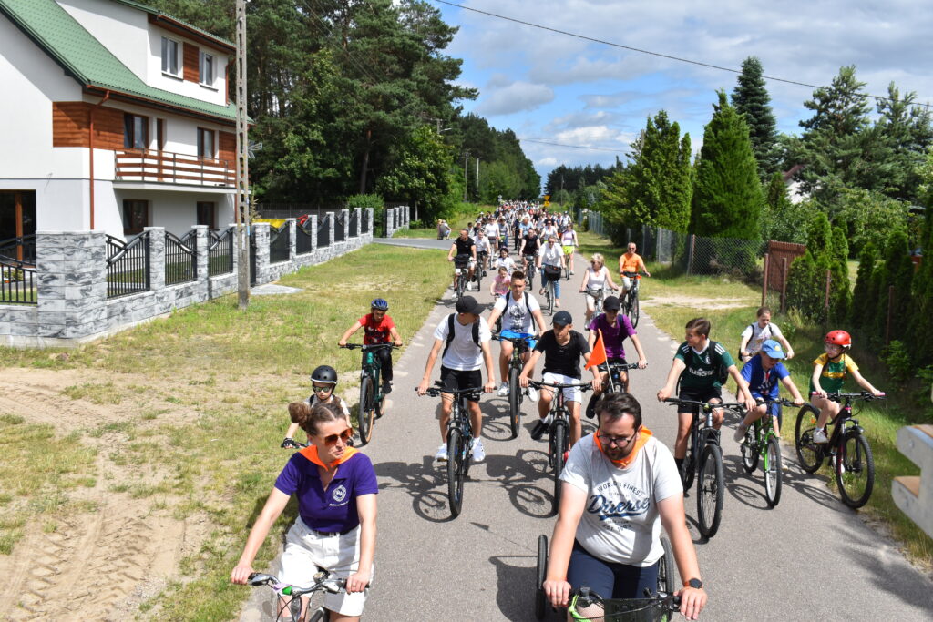 Ulicą w lesie jedzie grupa rowerzystów w kolorowych ubraniach. W tle dom i zielone drzewa. Niebo jest częściowo zachmurzone.