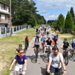 Ulicą w lesie jedzie grupa rowerzystów w kolorowych ubraniach. W tle dom i zielone drzewa. Niebo jest częściowo zachmurzone.