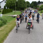 Na zdjęciu widoczna jest grupa rowerzystów na wiejskiej drodze. Otoczenie to zielone pola i drzewa, w tle domy. Ludzie mają kolorowe stroje, a pogoda jest słoneczna.