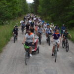 Grupa rowerzystów na leśnej ścieżce, otoczonej zielenią. Uczestnicy w kolorowych kaskach i strojach. Pogoda zachmurzona, tło z drzewami.