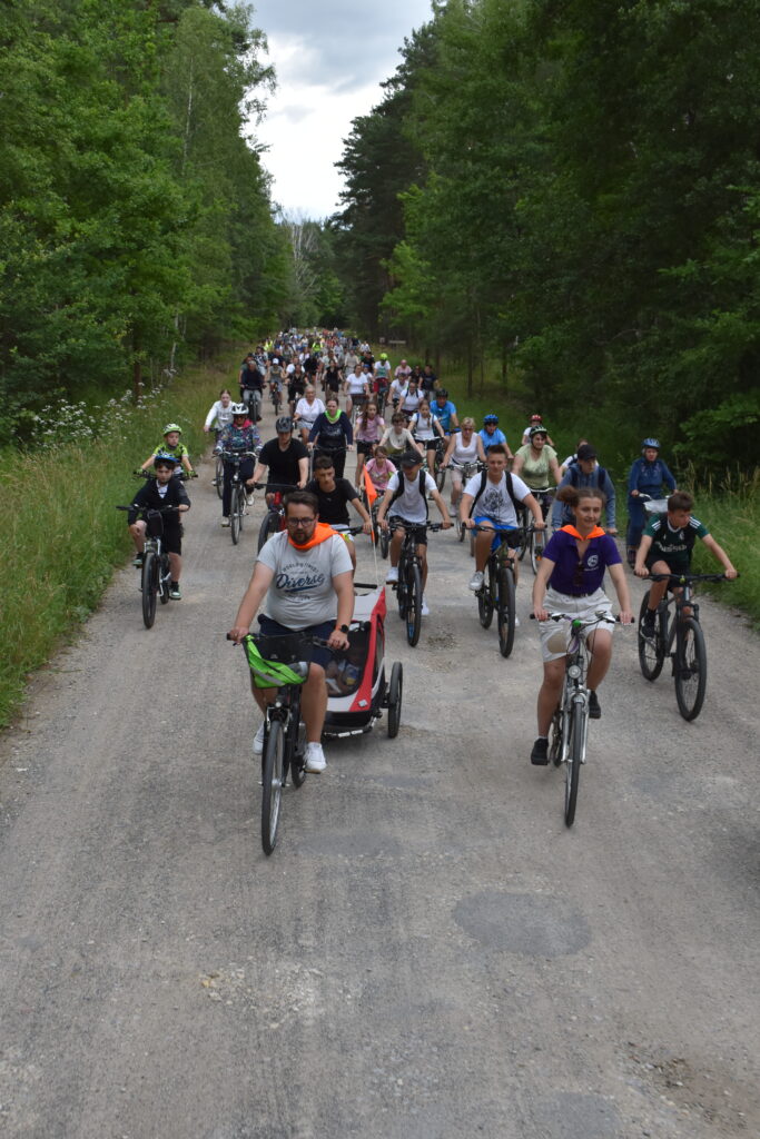 Grupa rowerzystów na leśnej ścieżce, otoczonej zielenią. Uczestnicy w kolorowych kaskach i strojach. Pogoda zachmurzona, tło z drzewami.