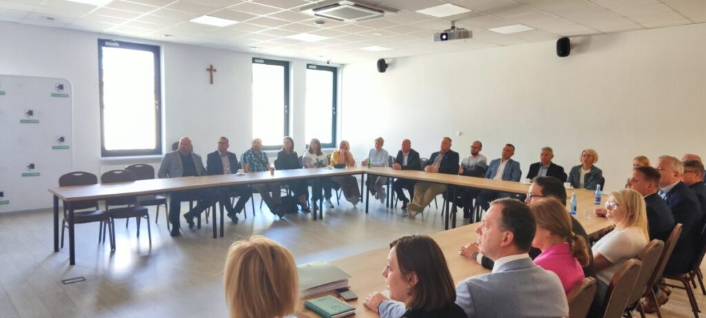 Zdjęcie przedstawia grupę ludzi siedzących przy dużym prostokątnym stole w jasnym pomieszczeniu. Na ścianie w tle widać krzyż i okna z jasnym światłem. Osoby noszą formalne ubrania, a oczy są zwrócone w jednym kierunku.