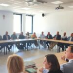 Zdjęcie przedstawia grupę ludzi siedzących przy dużym prostokątnym stole w jasnym pomieszczeniu. Na ścianie w tle widać krzyż i okna z jasnym światłem. Osoby noszą formalne ubrania, a oczy są zwrócone w jednym kierunku.