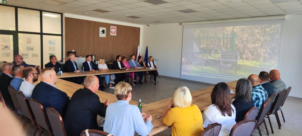 Sala konferencyjna z osobami siedzącymi przy stole w kształcie litery U. Na ścianie wyświetlany obraz z zielonym płotem. Ściany w jasnych kolorach, flaga w tle.