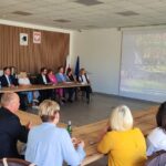 Sala konferencyjna z osobami siedzącymi przy stole w kształcie litery U. Na ścianie wyświetlany obraz z zielonym płotem. Ściany w jasnych kolorach, flaga w tle.