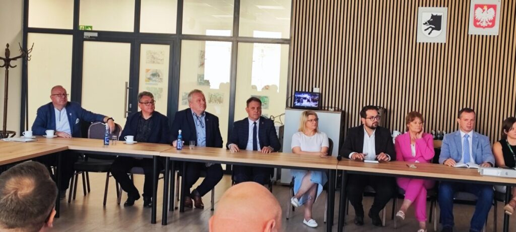 Grupa ludzi siedzi za długim stołem w sali konferencyjnej. Tło z drewnianymi panelami, godła na ścianie. Elegancka atmosfera spotkania.