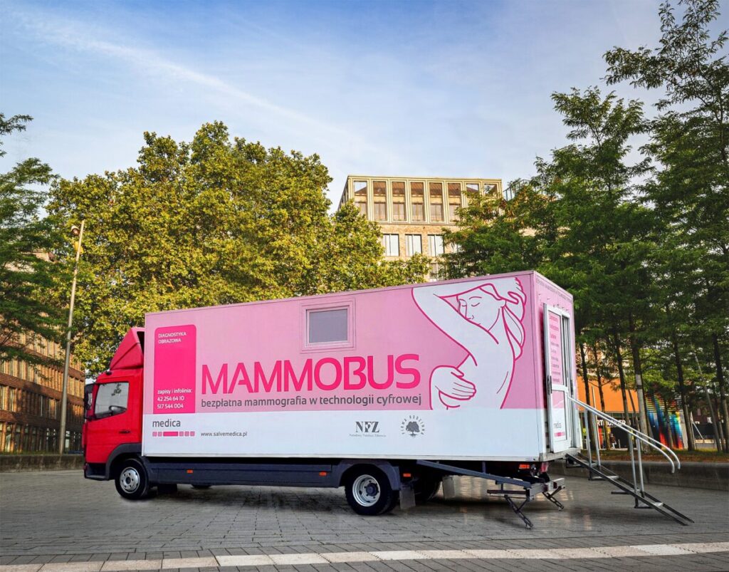Czerwony mammo-bus z różowym napisem stoi na placu. W tle drzewa, budynek i jasne niebo. Oferuje darmową mammografię.