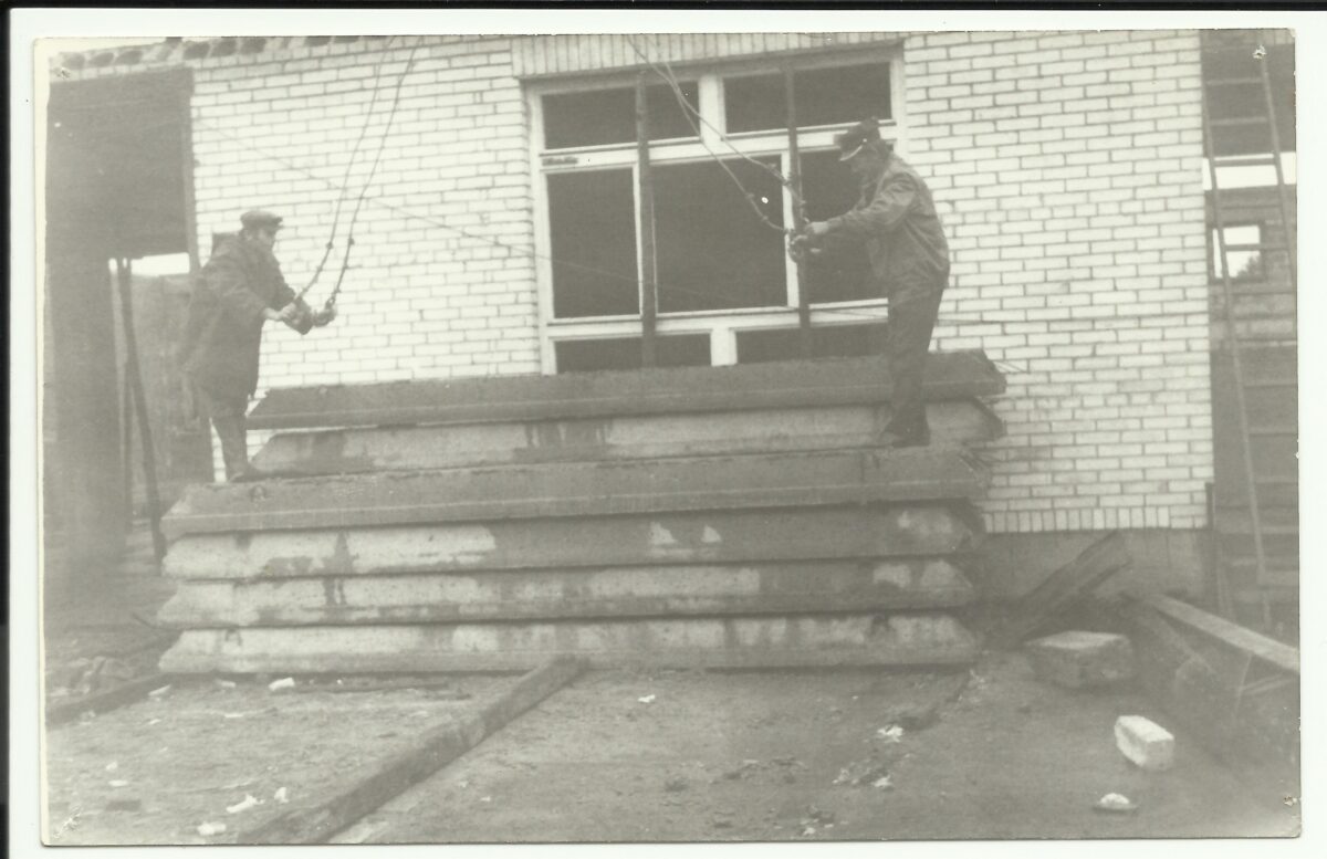 Dwóch mężczyzn pracuje na stosie betonowych płyt przed ceglaną ścianą. Obaj trzymają liny. Tło jest monochromatyczne.