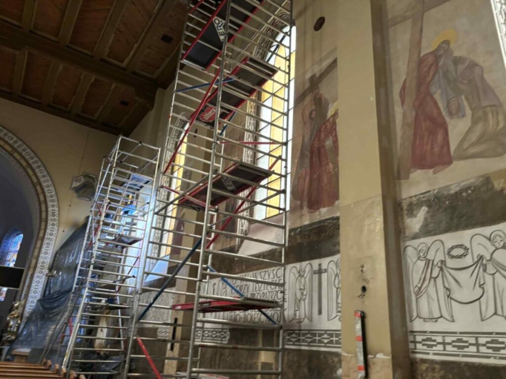Wnętrze kościoła z freskami na ścianach w odcieniach brązu i szarości. Na pierwszym planie stoją metalowe rusztowania. Na suficie widoczne drewniane belki.