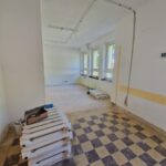 Pomieszczenie w remoncie; podłoga w żółto-brązową szachownicę, białe ściany. Zestawy kaloryferów ułożone na podłodze.