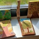 Na parapecie stoją malowane kawałki drewna z motywami drzew i przyrody. Przewaga zieleni, brązu i niebieskiego tła. Za oknem widać trawę.