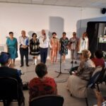 Na scenie stoi osiem osób, ubranych w różnorodne stroje. Tło jest jasne z zawieszonymi kolorowymi obrazami po prawej stronie. Publiczność siedzi na krzesłach, patrząc na występ.