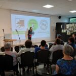 Spotkanie w sali z białymi ścianami, rzutnikiem i publicznością. Na ekranie logo i napisy, ludzie siedzą na krzesłach. Kobieta prezentuje coś przy mikrofonie.