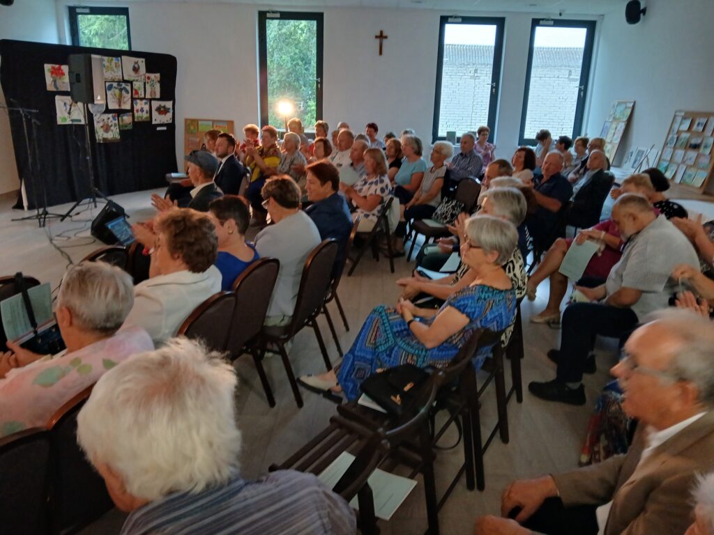 Grupa osób siedzi w jasnej sali, patrząc naprzód. Tło zdobią kolorowe prace na czarnych panelach. Na ścianie duże okna i krzyż.