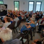 Grupa osób siedzi w jasnej sali, patrząc naprzód. Tło zdobią kolorowe prace na czarnych panelach. Na ścianie duże okna i krzyż.