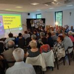 Spotkanie w sali z publicznością, projektor wyświetla napis „Uniwersytet Dobrego Wieku”. Przeważają jasne kolory. Dookoła są obrazy i krzyż na ścianie.