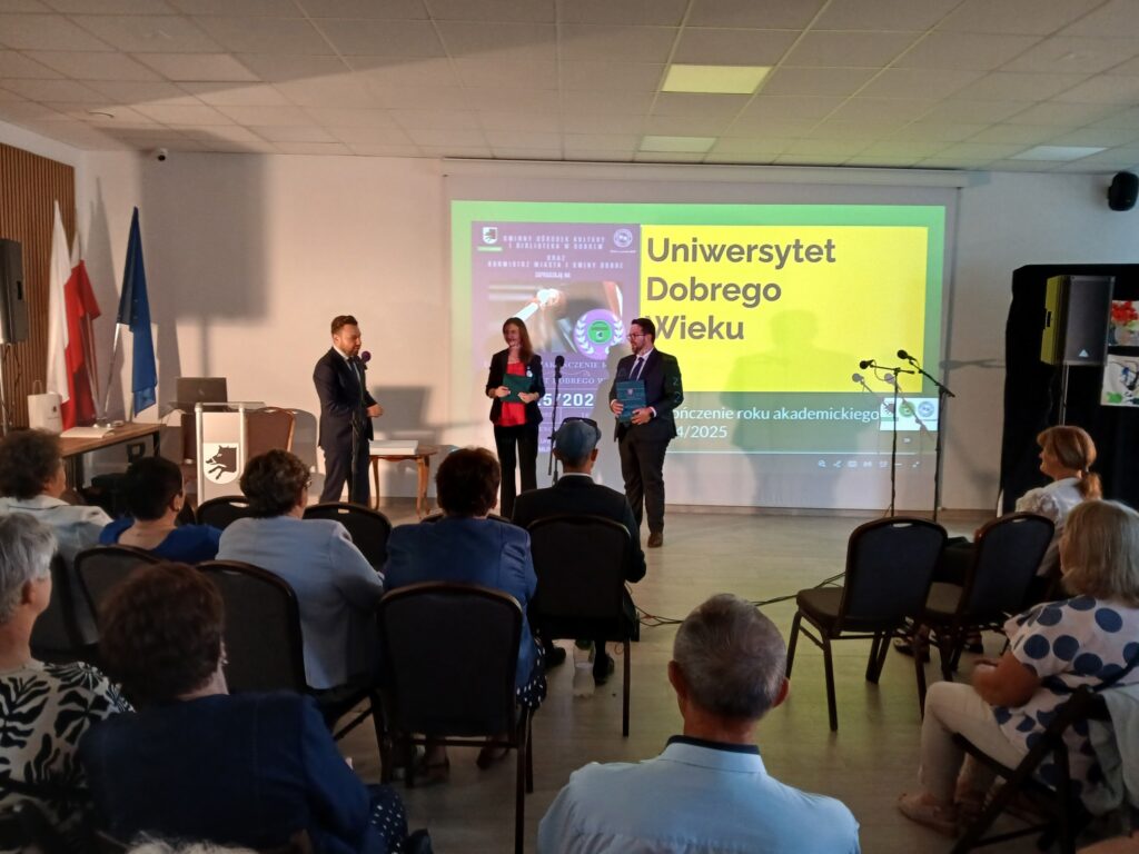 Starsze osoby siedzą w sali, patrząc na scenę, gdzie troje prelegentów stoi przy żółtym ekranie z napisem Uniwersytet Dobrego Wieku. Na ścianie widoczne są flagi i projektor. Sala jest jasna, z białymi ścianami
