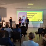 Starsze osoby siedzą w sali, patrząc na scenę, gdzie troje prelegentów stoi przy żółtym ekranie z napisem Uniwersytet Dobrego Wieku. Na ścianie widoczne są flagi i projektor. Sala jest jasna, z białymi ścianami