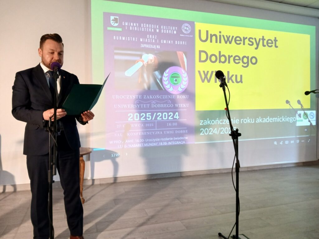 Na zdjęciu mężczyzna w garniturze przemawia do mikrofonu. Obok niego widoczna jest jasna ściana z zielono-żółtym ekranem z napisem Uniwersytet Dobrego Wieku. Pomieszczenie jest dobrze oświetlone, a tło jest neutralne.