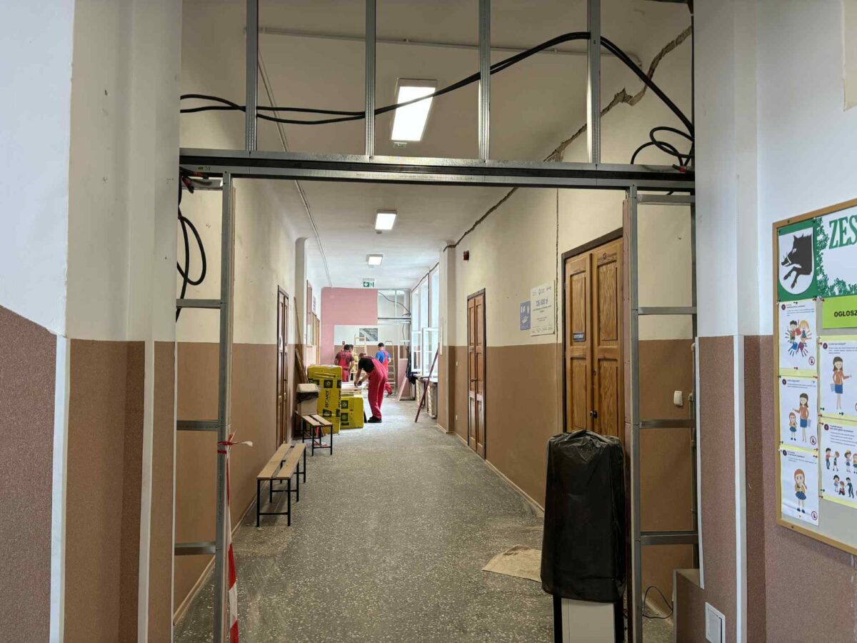 Korytarz szkolny w trakcie remontu, z pracownikami i narzędziami. Ściany biało-brązowe, podłoga szara. Widoczne oznaki napraw.