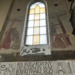 Na obrazie widać fresk z ukrzyżowaniem, utrzymany w stonowanych kolorach. Na tle są jasne witraże okienne, wpuszczające światło. Dół fresku zdobią anioły oraz inskrypcja religijna.